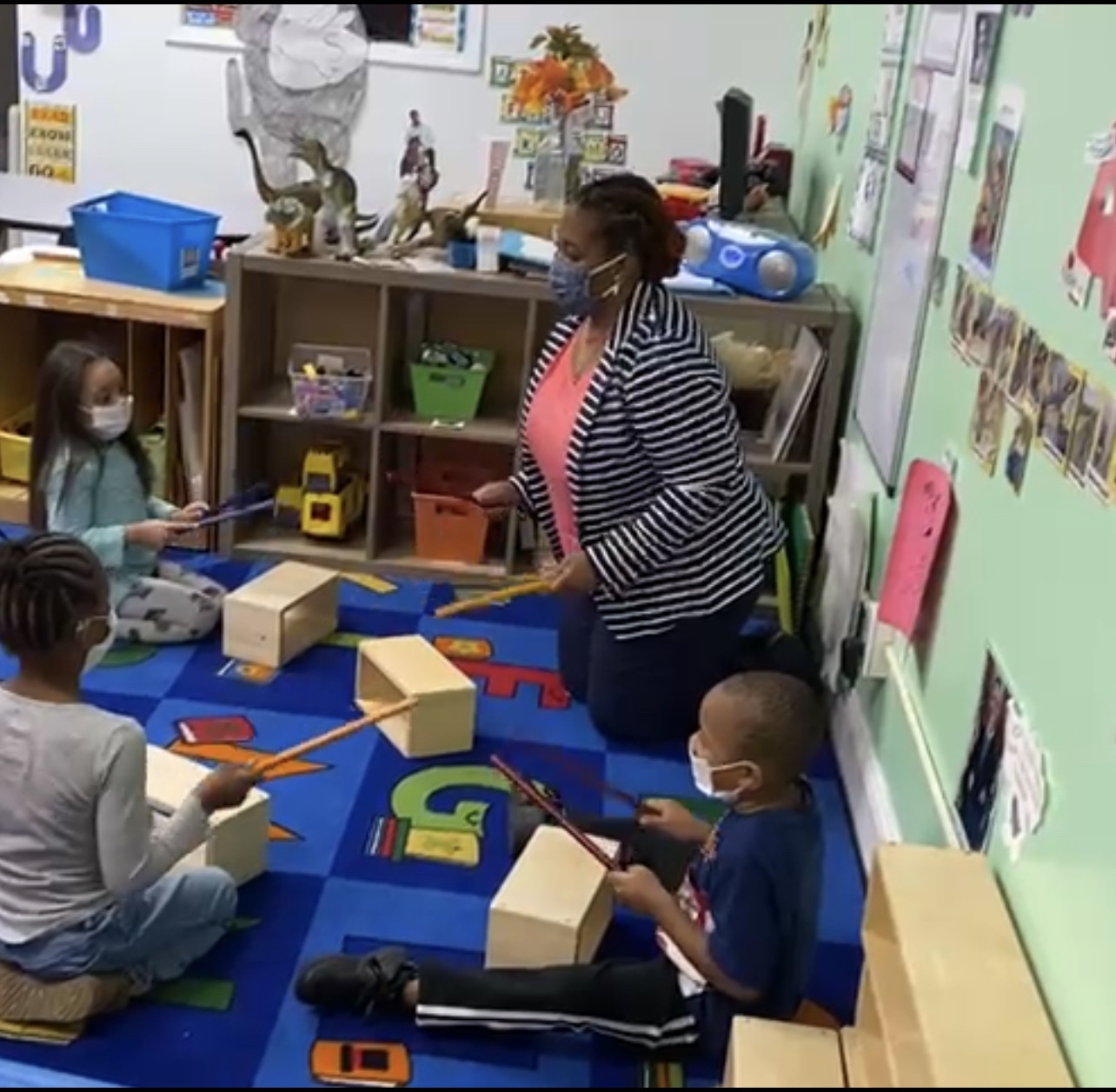A small group of children sit in a circle around a teacher exploring making sounds with blocks and wooden sticks.