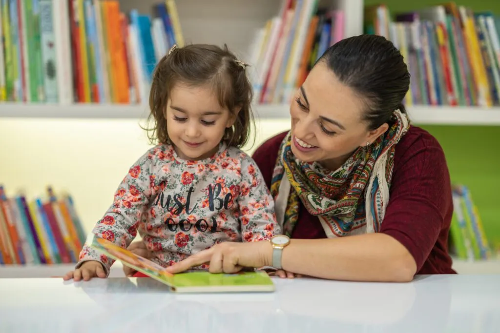 An adult woman sits one-on-one with a toddler at a table in a library, pointing to illustrations in a board book and talking about them together.